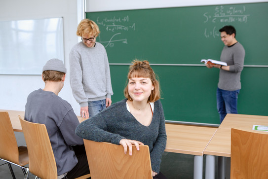Vier Studierende in einem Klassenzimmer. Ein Student steht mit einem Buch vor einer grünen Tafel auf der mathematische Formeln zu sehen sind. Zwei weitere Studenten wirken im Gespräch versunken, während eine rothaarige, junge Studentin sich auf dem Stuhl zum Betrachter dreht und herausfordernd in die Kamera lächelt.