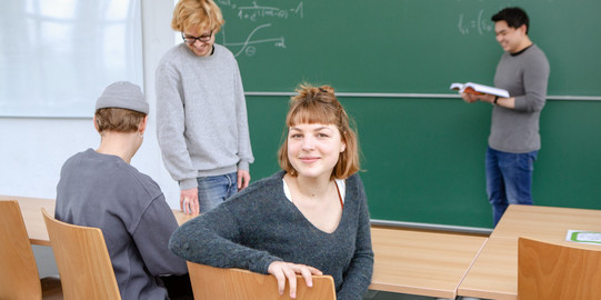 Vier Studierende in einem Klassenzimmer. Ein Student steht mit einem Buch vor einer grünen Tafel auf der mathematische Formeln zu sehen sind. Zwei weitere Studenten wirken im Gespräch versunken, während eine rothaarige, junge Studentin sich auf dem Stuhl zum Betrachter dreht und herausfordernd in die Kamera lächelt.